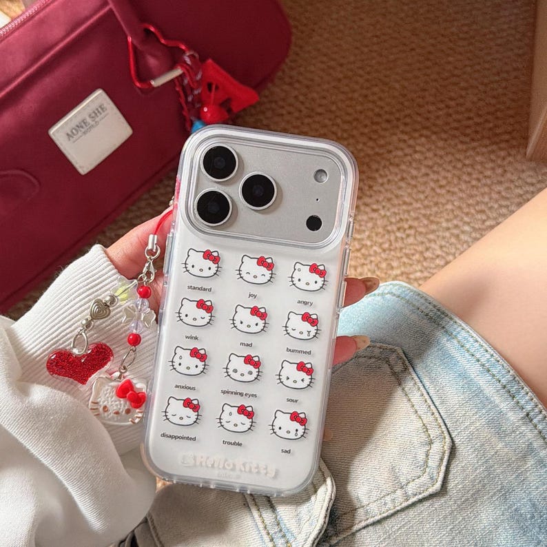K&ouml;nnte beinhalten: Transparente Handyh&uuml;lle mit einem sich wiederholenden Muster von Hello Kitty-Gesichtern mit verschiedenen Ausdr&uuml;cken. Die H&uuml;lle hat ein silbernes Kameramodul und den Schriftzug "Hello Kitty". Eine rote Tasche und ein Herz-Charm sind ebenfalls zu sehen.