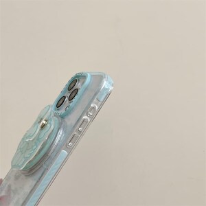 K&ouml;nnte beinhalten: Transparente Handyh&uuml;lle mit hellblauem Akzent um die Kamera und die Kanten. Die H&uuml;lle hat einen hellblauen Pop-up-Griff mit floralem Design. Die Handyh&uuml;lle dient zum Schutz des Telefons und bietet einen sicheren Halt.