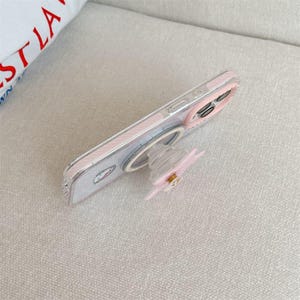K&ouml;nnte beinhalten: Eine transparente Handyh&uuml;lle mit rosa und wei&szlig;em Design, mit einem Pop-up-Griff mit einem rosa Blumenmuster. Die H&uuml;lle hat einen Ringhalter und eine kleine Grafik auf der R&uuml;ckseite.