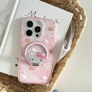 May include: Pink phone case featuring a Hello Kitty design with a matching ring holder. The case has a clear border and is decorated with stars and Hello Kitty faces. The phone case is resting in a woven basket.