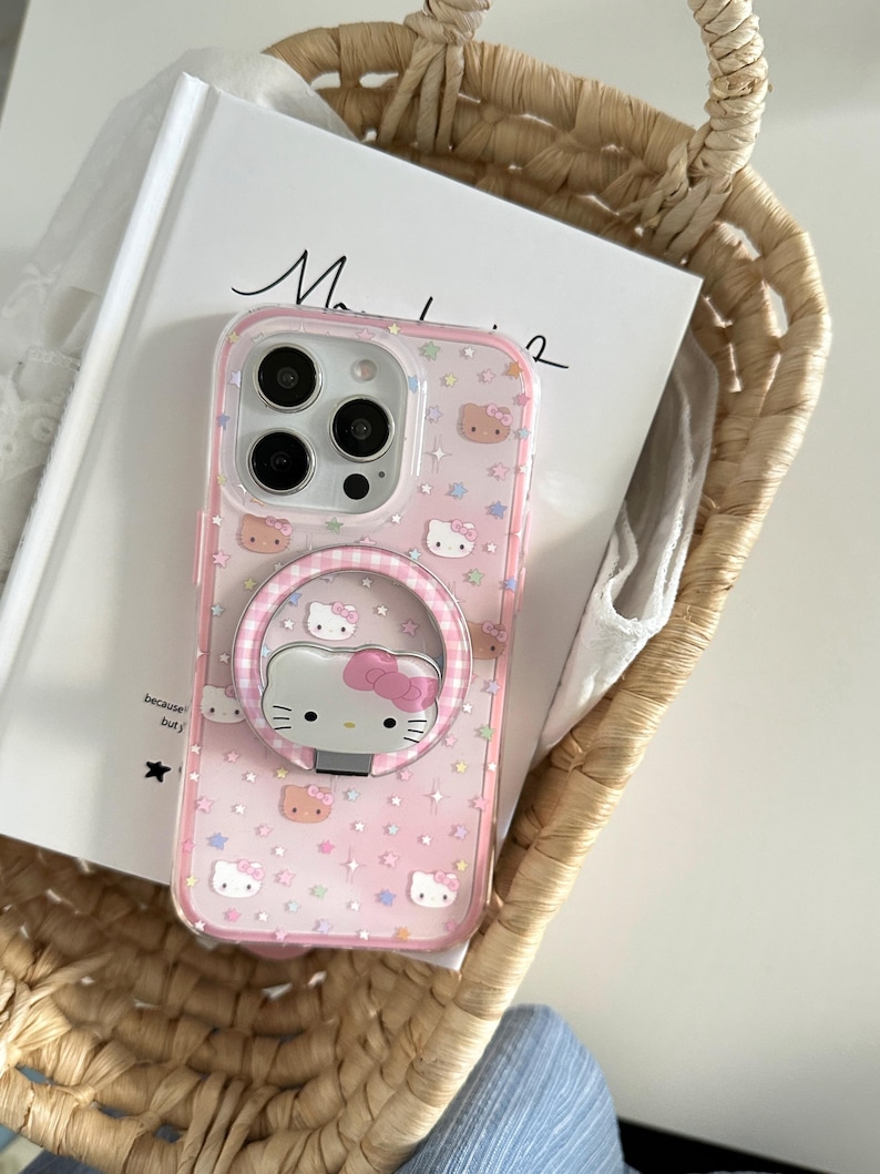 May include: Pink phone case featuring a Hello Kitty design with a matching ring holder. The case has a clear border and is decorated with stars and Hello Kitty faces. The phone case is resting in a woven basket.