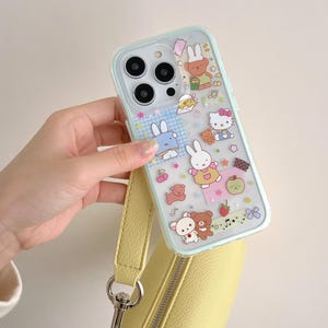 May include: A clear phone case with a light blue border featuring cartoon characters, including bunnies, bears, and Hello Kitty. The case is held by a yellow strap attached to a yellow purse.