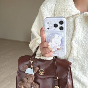 Op de afbeelding: Een doorzichtige telefoonhoes met een cartoon konijn ontwerp wordt getoond. De hoes heeft een wit konijn met een roze jurk en een bijpassende konijn charme. Een bruine tas met een teddybeer sleutelhanger hangt eronder.