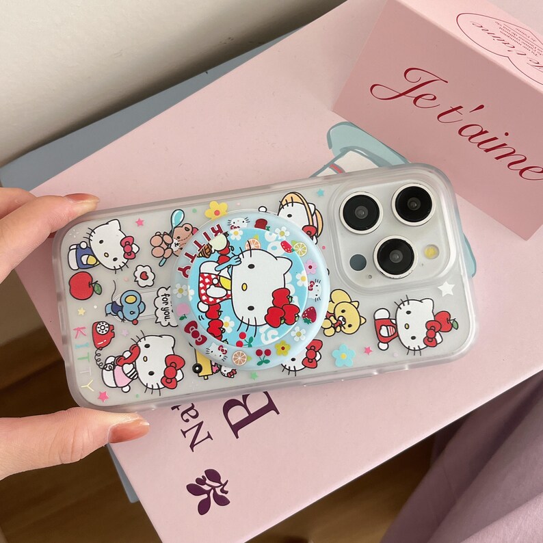 May include: Clear phone case with a colorful Hello Kitty design. The case features a pop-up grip with a Hello Kitty graphic and is decorated with various Hello Kitty characters, flowers, and stars. The case is on a pink surface.