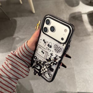 Op de afbeelding: Een zwarte en doorzichtige telefoonhoes met een decoratief ontwerp met een verscheidenheid aan kleine illustraties, waaronder een kat, een handtas en een hart. De hoes heeft een zwart-witte kralenbedel.