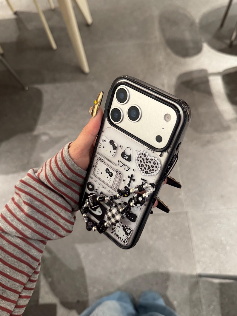 Op de afbeelding: Een zwarte en doorzichtige telefoonhoes met een decoratief ontwerp met een verscheidenheid aan kleine illustraties, waaronder een kat, een handtas en een hart. De hoes heeft een zwart-witte kralenbedel.