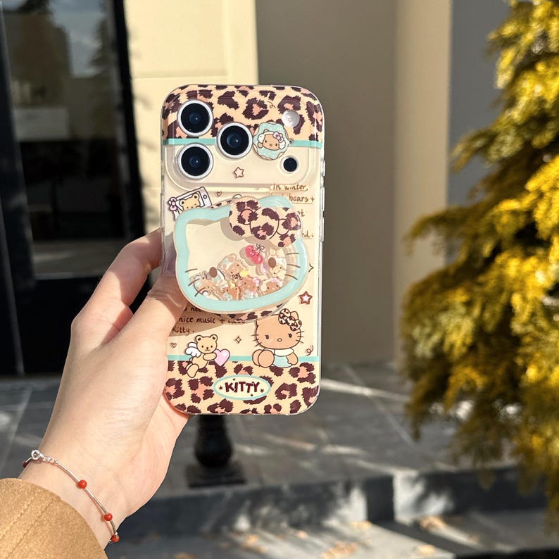 May include: A phone case with a leopard print design and a clear, liquid-filled section shaped like a cat's head. The case features cartoon illustrations of a bear, a cat, and the word "Kitty". The case has a built-in phone grip.