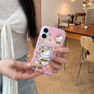 May include: Pink phone case featuring Hello Kitty and bee-themed designs. The case has a clear ring holder with a Hello Kitty graphic and a pink flower. The case is decorated with small flower and star shapes.