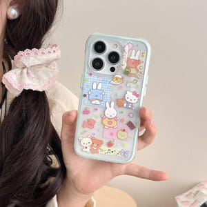 May include: A clear phone case with a light blue border, decorated with cartoon characters including bunnies, bears, and Hello Kitty. The case has a variety of colourful stickers and designs, including stars, strawberries, and musical notes.