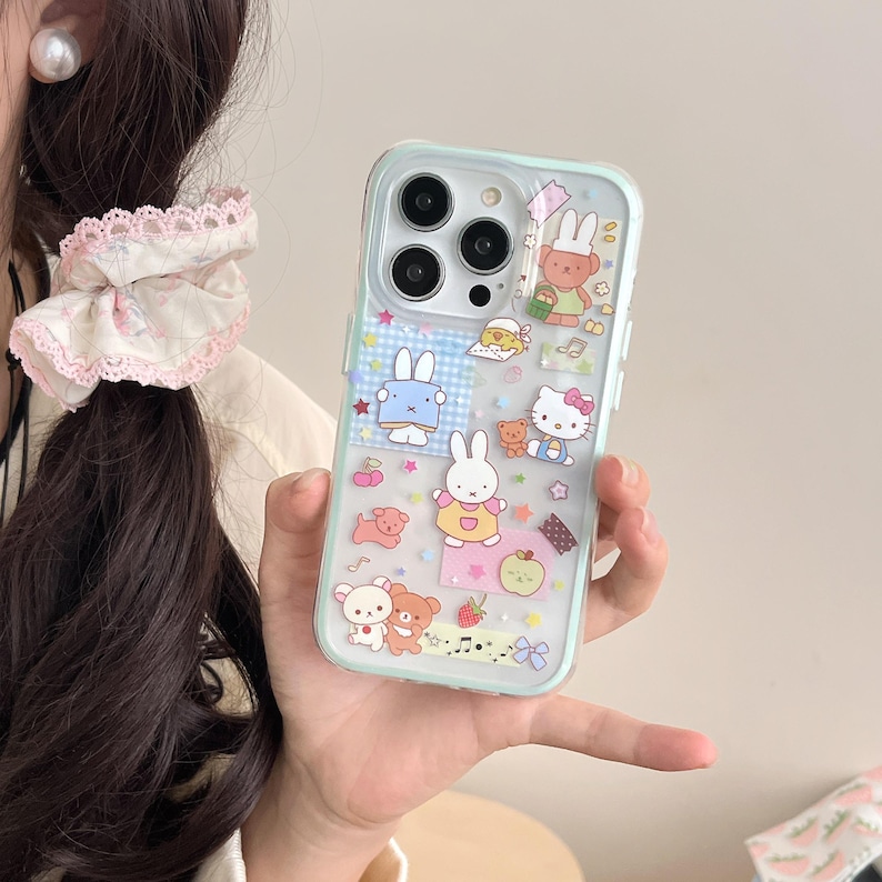 May include: A clear phone case with a light blue border, decorated with cartoon characters including bunnies, bears, and Hello Kitty. The case has a variety of colourful stickers and designs, including stars, strawberries, and musical notes.