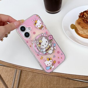 May include: Pink phone case featuring Hello Kitty characters dressed as bees, with musical notes and flower accents. The case includes a clear ring holder with a Hello Kitty design and a clear border.