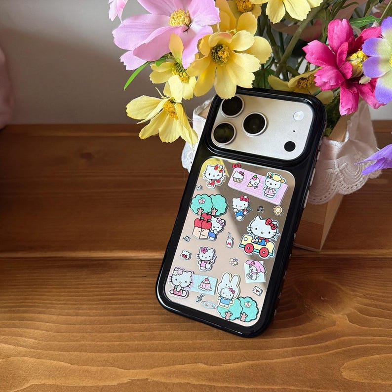 May include: Black phone case with a clear center featuring Hello Kitty and other cartoon character stickers. The case has a black border and a clear back, showcasing the phone's camera and flash. The background includes colorful flowers.