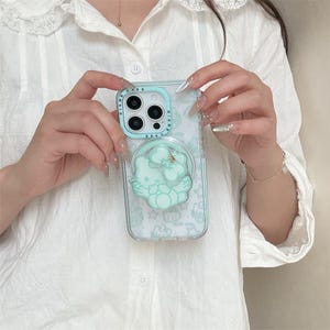 K&ouml;nnte beinhalten: Transparente Handyh&uuml;lle mit hellblauem Sto&szlig;f&auml;nger und hellblauem Hello Kitty-Design. Die H&uuml;lle hat einen Pop-up-Griff mit einer goldenen Schleife. Die Handyh&uuml;lle wird vor einem wei&szlig;en Hemd gehalten.