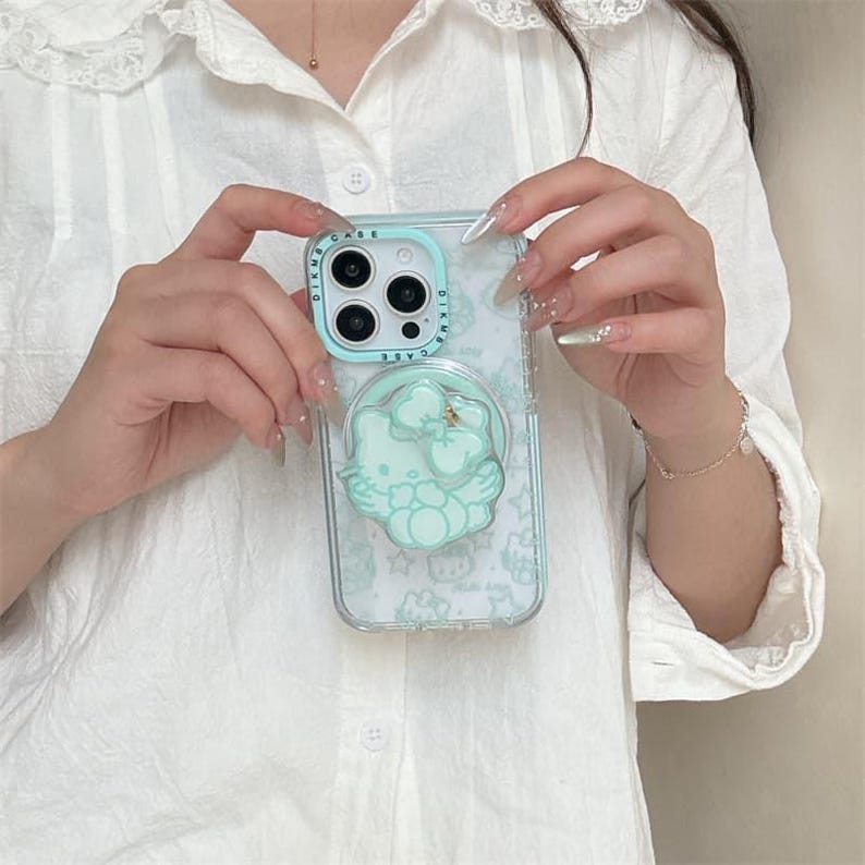 K&ouml;nnte beinhalten: Transparente Handyh&uuml;lle mit hellblauem Sto&szlig;f&auml;nger und hellblauem Hello Kitty-Design. Die H&uuml;lle hat einen Pop-up-Griff mit einer goldenen Schleife. Die Handyh&uuml;lle wird vor einem wei&szlig;en Hemd gehalten.