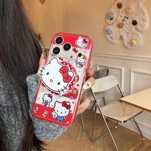 May include: Red and clear phone case featuring Hello Kitty designs. The case has a pop-up grip with a large Hello Kitty face and a red bow. The case is decorated with various Hello Kitty illustrations and a red border.