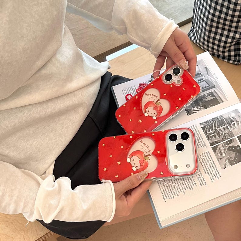 Puede incluir: Dos fundas rojas para tel&eacute;fono con un dise&ntilde;o de Hello Kitty. Cada funda presenta un coraz&oacute;n blanco con Hello Kitty como manzana, rodeado de estrellas doradas. Las fundas se sostienen sobre un libro abierto.