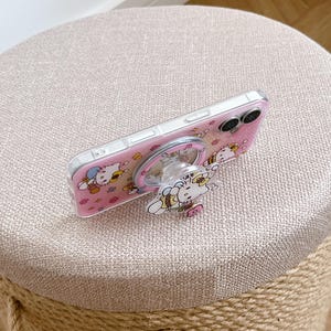 May include: A clear phone case with a pink background featuring a cartoon cat design. The case has a matching pop-up grip with the same cat design. The phone case is resting on a beige, round stool.