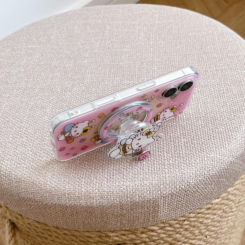 May include: A clear phone case with a pink background featuring a cartoon cat design. The case has a matching pop-up grip with the same cat design. The phone case is resting on a beige, round stool.