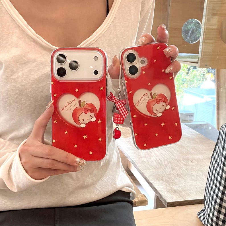 Puede incluir: Dos fundas rojas para tel&eacute;fono con un dise&ntilde;o de Hello Kitty. Cada funda presenta un recorte en forma de coraz&oacute;n con una ilustraci&oacute;n de manzana y Hello Kitty, rodeada de peque&ntilde;as estrellas blancas. Una funda tiene una cinta a cuadros roja y blanca y un peque&ntilde;o dije rojo.