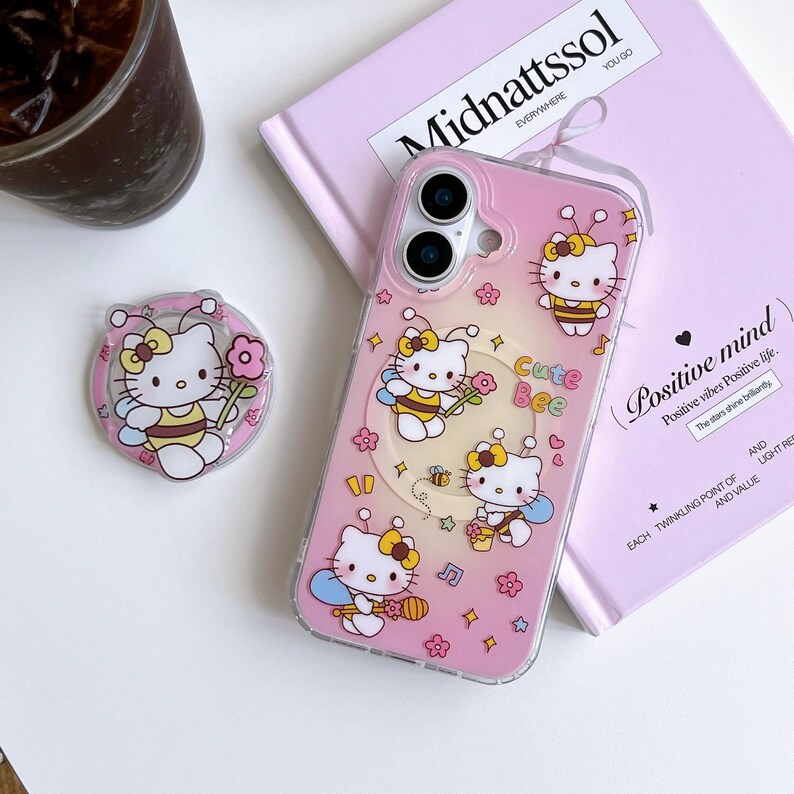 May include: A pink phone case and a matching phone grip featuring a cartoon cat dressed as a bee. The case has the words "Cute Bee" and "Positive mind" printed on it. The phone grip is round with a pink border.