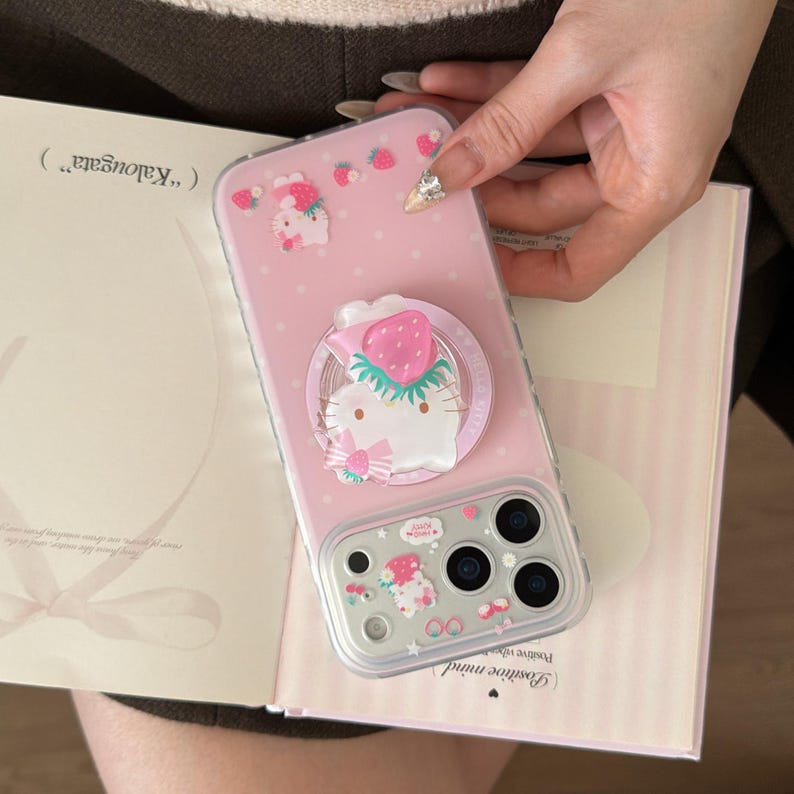 May include: Pink phone case with a strawberry and Hello Kitty design. The case has a pop-up grip with a strawberry on top. The case is decorated with small strawberry and Hello Kitty graphics.