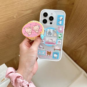 Puede incluir: Funda transparente para tel&eacute;fono con una variedad de pegatinas de Hello Kitty. Un pop socket rosa y amarillo con dos personajes de Hello Kitty est&aacute; adjunto. Un dije azul claro cuelga del pop socket.