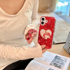 Puede incluir: Funda de tel&eacute;fono roja con borde transparente, con un dise&ntilde;o en forma de coraz&oacute;n con un gr&aacute;fico de manzana Hello Kitty y estrellas blancas. Un llavero de lazo rojo y blanco est&aacute; adjunto. La funda del tel&eacute;fono se sostiene sobre un libro abierto.