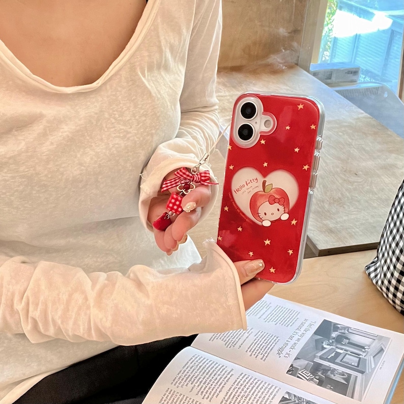 Puede incluir: Funda de tel&eacute;fono roja con borde transparente, con un dise&ntilde;o en forma de coraz&oacute;n con un gr&aacute;fico de manzana Hello Kitty y estrellas blancas. Un llavero de lazo rojo y blanco est&aacute; adjunto. La funda del tel&eacute;fono se sostiene sobre un libro abierto.