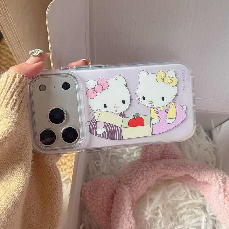 May include: Clear phone case featuring a cartoon illustration of two Hello Kitty characters. One Kitty wears a pink bow and the other a yellow bow. The case has a light purple background and a camera cutout.