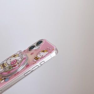 May include: A pink phone case with a clear, transparent bumper. The case features a cartoon design of a cat with a yellow bow and a bee. A clear phone grip with a floral design is attached to the case.