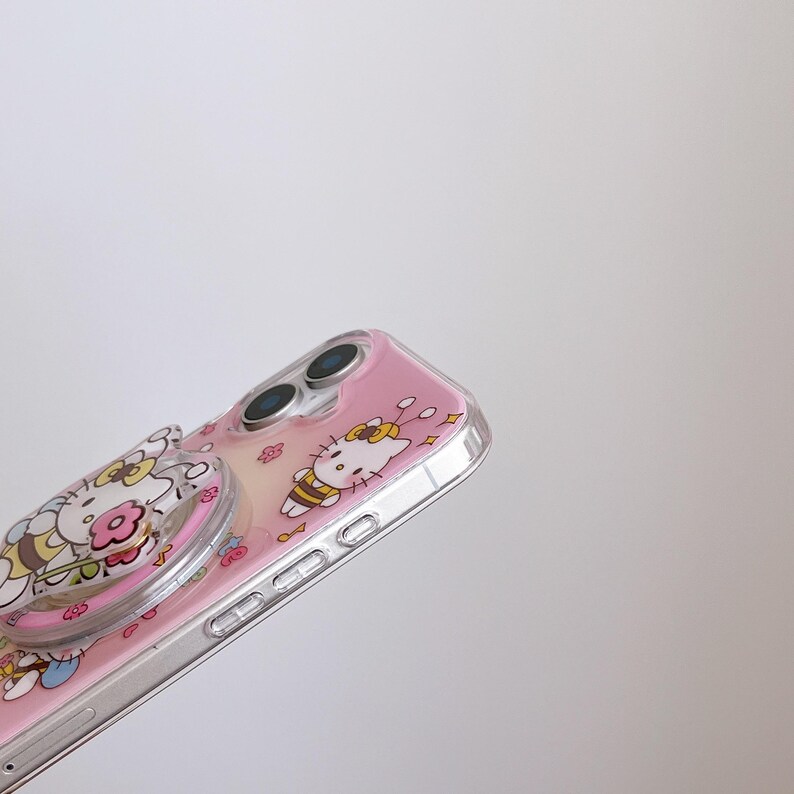 May include: A pink phone case with a clear, transparent bumper. The case features a cartoon design of a cat with a yellow bow and a bee. A clear phone grip with a floral design is attached to the case.