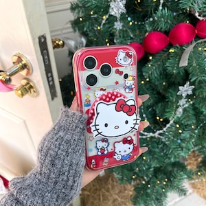 May include: A red and clear phone case featuring Hello Kitty designs. The case has a red border and a pop-up grip with a Hello Kitty face. The design includes multiple Hello Kitty characters, bows, and stars. The case is designed to fit a smartphone.
