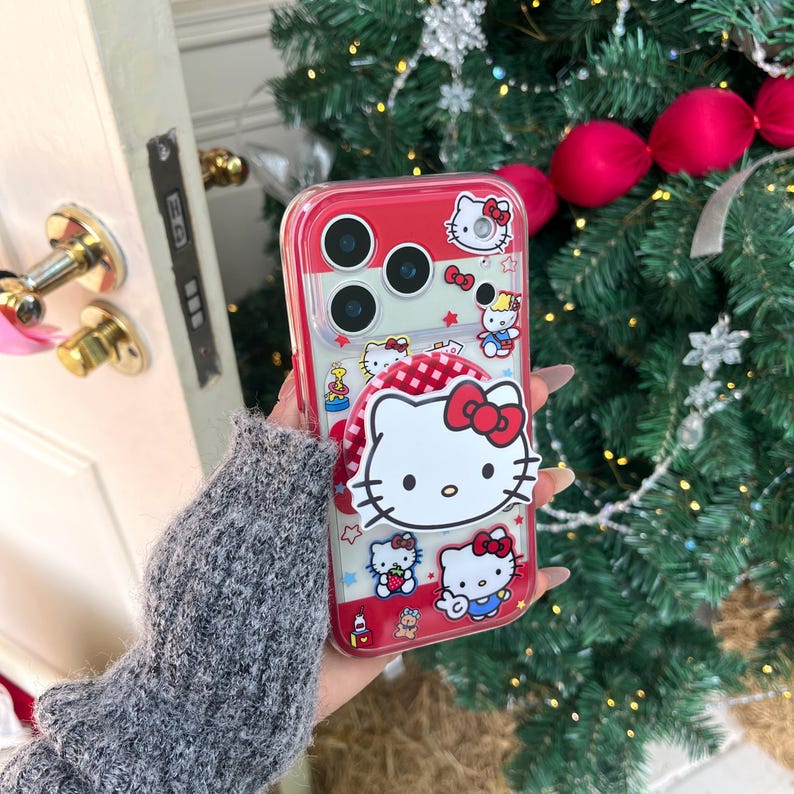 May include: A red and clear phone case featuring Hello Kitty designs. The case has a red border and a pop-up grip with a Hello Kitty face. The design includes multiple Hello Kitty characters, bows, and stars. The case is designed to fit a smartphone.