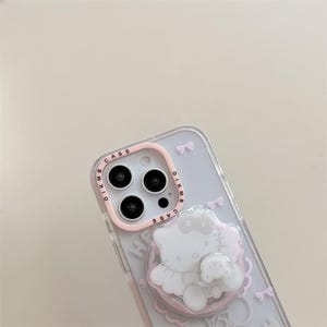 K&ouml;nnte beinhalten: Transparente Handyh&uuml;lle mit rosa Kameraring und 3D-Hello Kitty-Design. Die H&uuml;lle zeigt eine wei&szlig;e Hello Kitty-Figur mit Schleife, eine kleinere B&auml;renfigur und rosa Schleifenakzente. Der Kameraring tr&auml;gt den Text "DIKMB CASE".