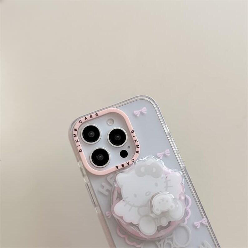 K&ouml;nnte beinhalten: Transparente Handyh&uuml;lle mit rosa Kameraring und 3D-Hello Kitty-Design. Die H&uuml;lle zeigt eine wei&szlig;e Hello Kitty-Figur mit Schleife, eine kleinere B&auml;renfigur und rosa Schleifenakzente. Der Kameraring tr&auml;gt den Text "DIKMB CASE".