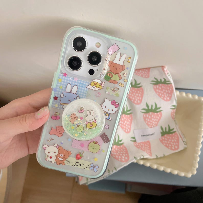 May include: A clear phone case with a mint green border, decorated with cartoon characters and a glittery pop socket. A white and pink strawberry-patterned cloth is next to the phone case.