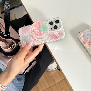 May include: Clear phone case with a pink and white design featuring cartoon characters and a strawberry pop socket. The case has a clear border and a pink background with small illustrations.