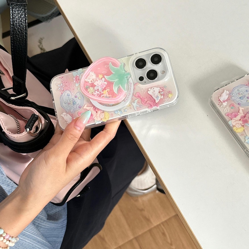 May include: Clear phone case with a pink and white design featuring cartoon characters and a strawberry pop socket. The case has a clear border and a pink background with small illustrations.