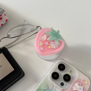 May include: A pink strawberry-shaped phone grip with a green leaf design. The grip features cartoon characters inside. A clear phone case with a similar design is also visible.