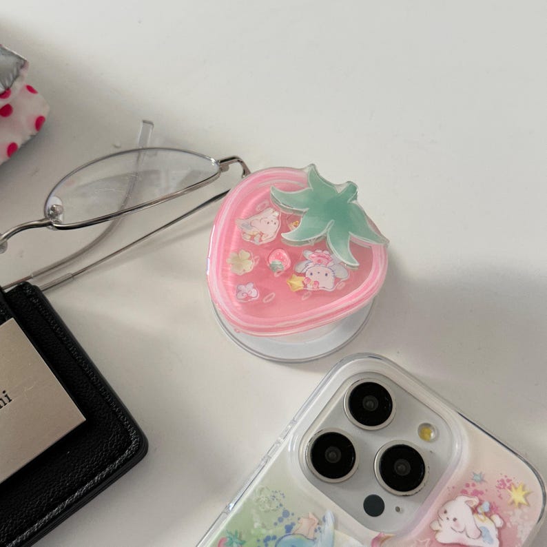 May include: A pink strawberry-shaped phone grip with a green leaf design. The grip features cartoon characters inside. A clear phone case with a similar design is also visible.