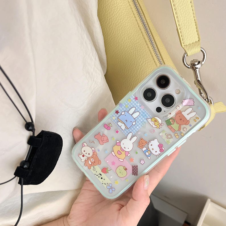 May include: A transparent phone case with a pale blue edge, decorated with cartoon images of rabbits, bears, and other characters. The case has a yellow strap attached and is held in a person's hand.