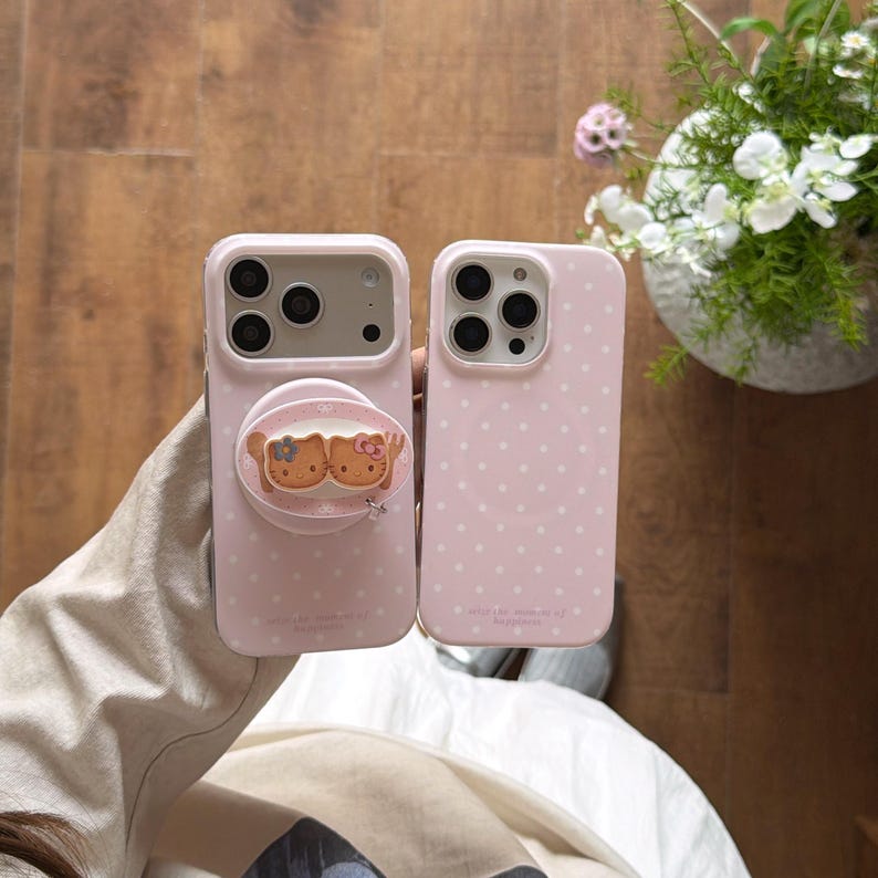May include: Two pink phone cases. One case has a pop-up grip with a cartoon design. The other case has a polka dot pattern. Both cases have camera cutouts. The cases are light pink.