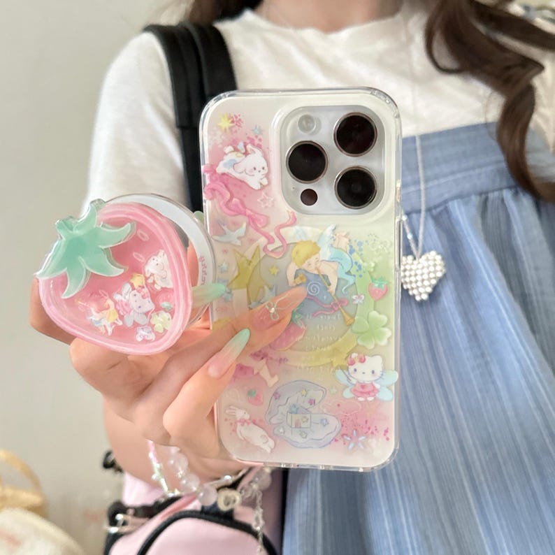 May include: Clear phone case with colorful illustrations of cartoon characters and a strawberry-shaped phone grip. The case features a variety of characters in a whimsical design. The strawberry grip is pink with a green stem.