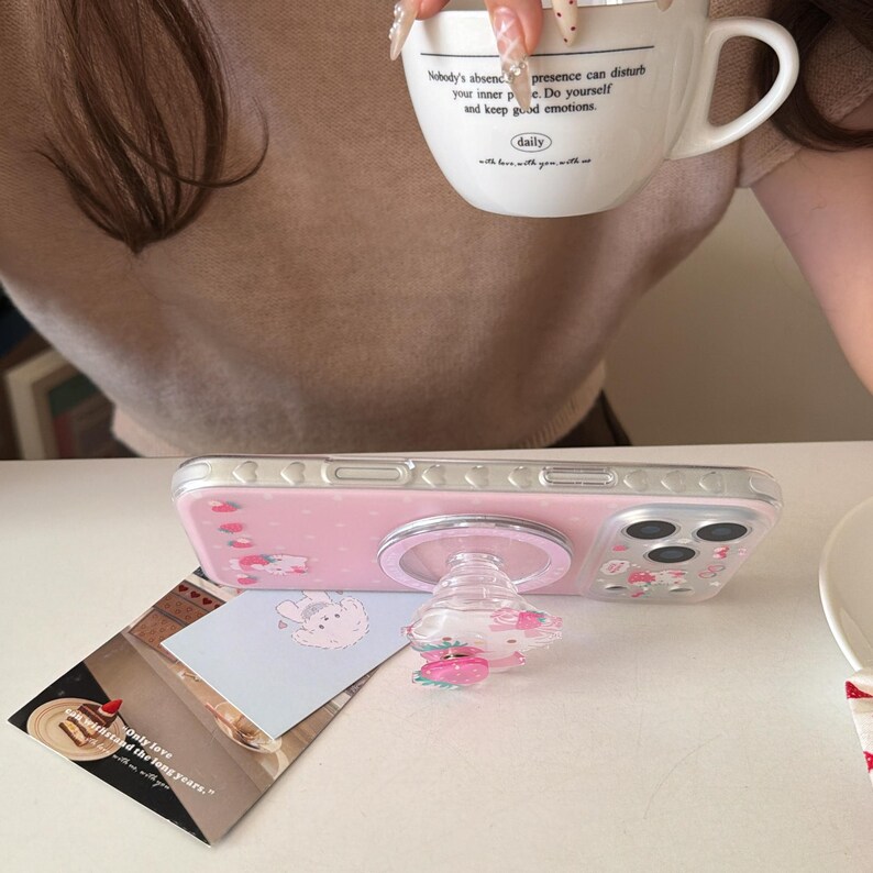 May include: A pink phone case with a clear pop socket featuring a strawberry design. The phone case has a pink background with cartoon illustrations. A white mug with text is held in the background.