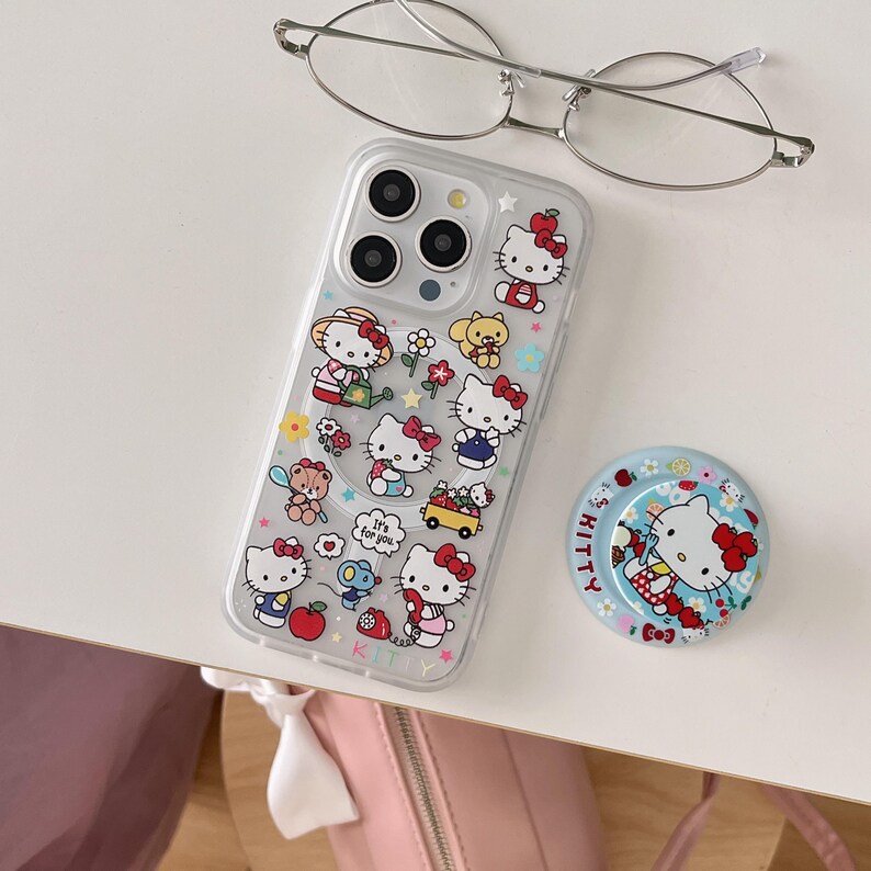 May include: Clear phone case with a Hello Kitty design, featuring various illustrations of the character with red bows, flowers, and other cute elements. A matching pop socket and eyeglasses are also present.