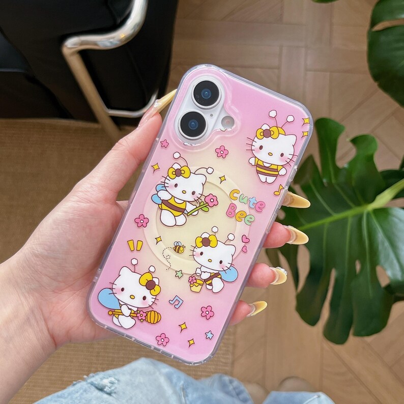 May include: Pink phone case featuring a cartoon design with Hello Kitty characters dressed as bees. The case has a clear border and includes the text "Cute Bee" along with floral and star accents. The phone case is designed to fit a smartphone.
