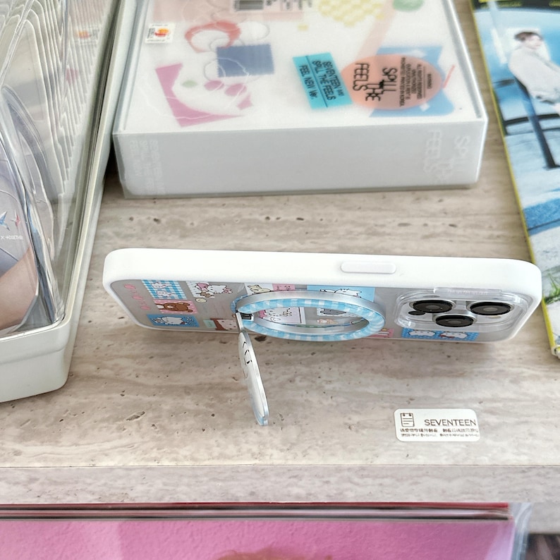 Puede incluir: Funda de tel&eacute;fono blanca con dise&ntilde;o de dibujos animados y agarre desplegable. La funda tiene una parte trasera transparente con un patr&oacute;n colorido y un agarre de anillo plateado. El agarre desplegable est&aacute; extendido.