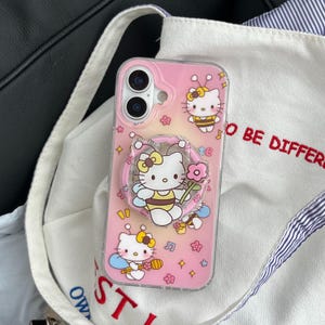 May include: Pink phone case featuring Hello Kitty dressed as a bee, holding a flower, and surrounded by musical notes and stars. The case has a pop-up grip with the text "be different".