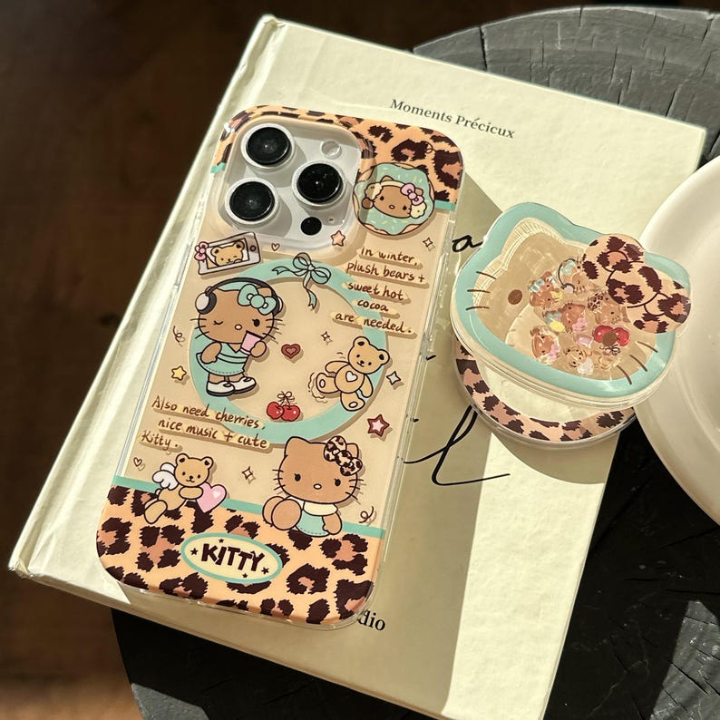 May include: Clear phone case with a leopard print design and illustrations of Hello Kitty characters. The case has a matching pop socket with a Hello Kitty design. Text on the case includes "Kitty" and other phrases.