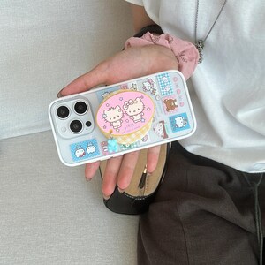 Puede incluir: Una funda de tel&eacute;fono de color claro con un dise&ntilde;o colorido de personajes de dibujos animados y sellos. La funda tiene un agarre emergente con dos gatos de dibujos animados y un dije de flor azul. La funda est&aacute; dise&ntilde;ada para un tel&eacute;fono inteligente.